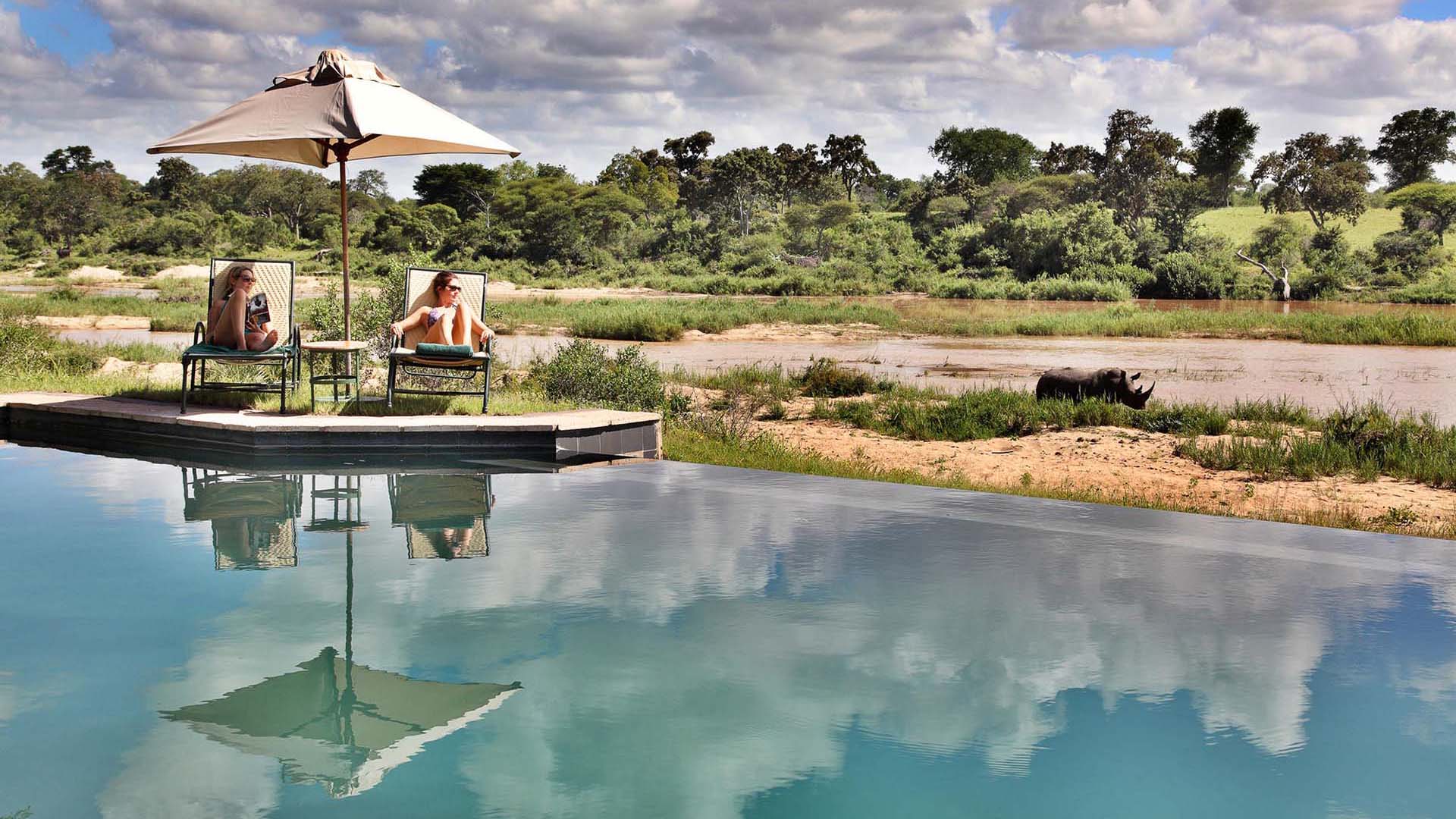2 ragazze ai bordi della piscina a Mala Mala osservano un rinoceronte che passeggia lungo il fiume